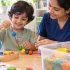 Young child participating in early childhood education programs through occupational therapy, threading colorful beads while a therapist provides gentle guidance in a bright child development center.